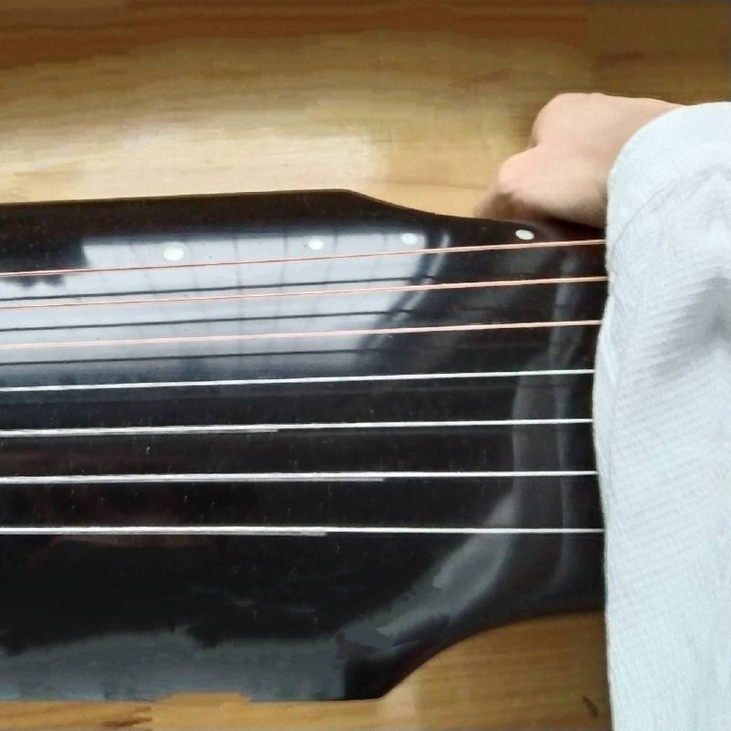 A hand in a light sleeve reaches across the strings of a glossy black zither-like instrument on a wooden surface.