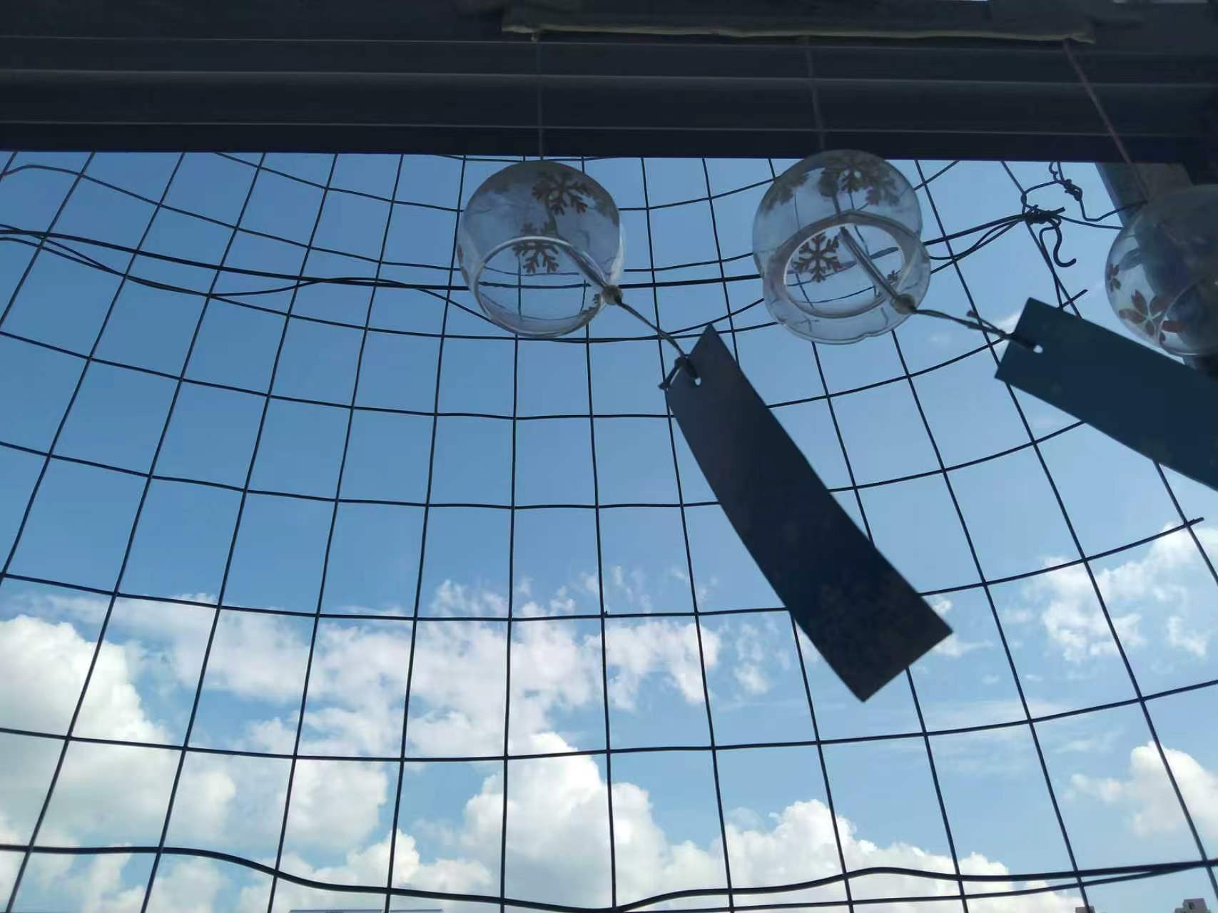 Glass wind chimes hanging from a wire grid against a blue sky with white clouds.