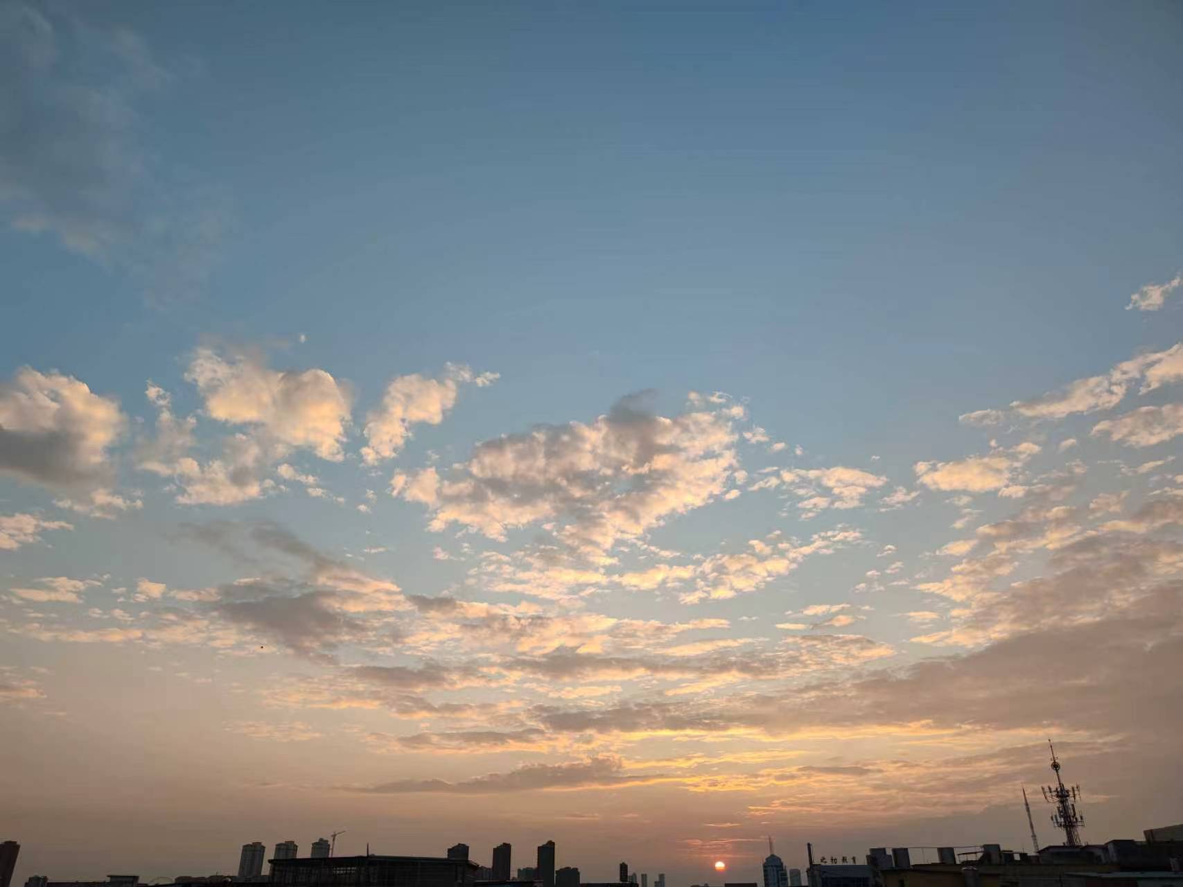 Sunset sky with scattered clouds, the sun near the horizon, and a dark city skyline below.
