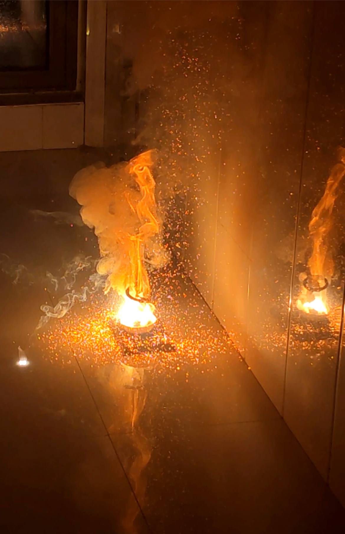 Indoor fireworks burn on a reflective floor, sending orange sparks and smoke upward with a bright reflection on the tiles.