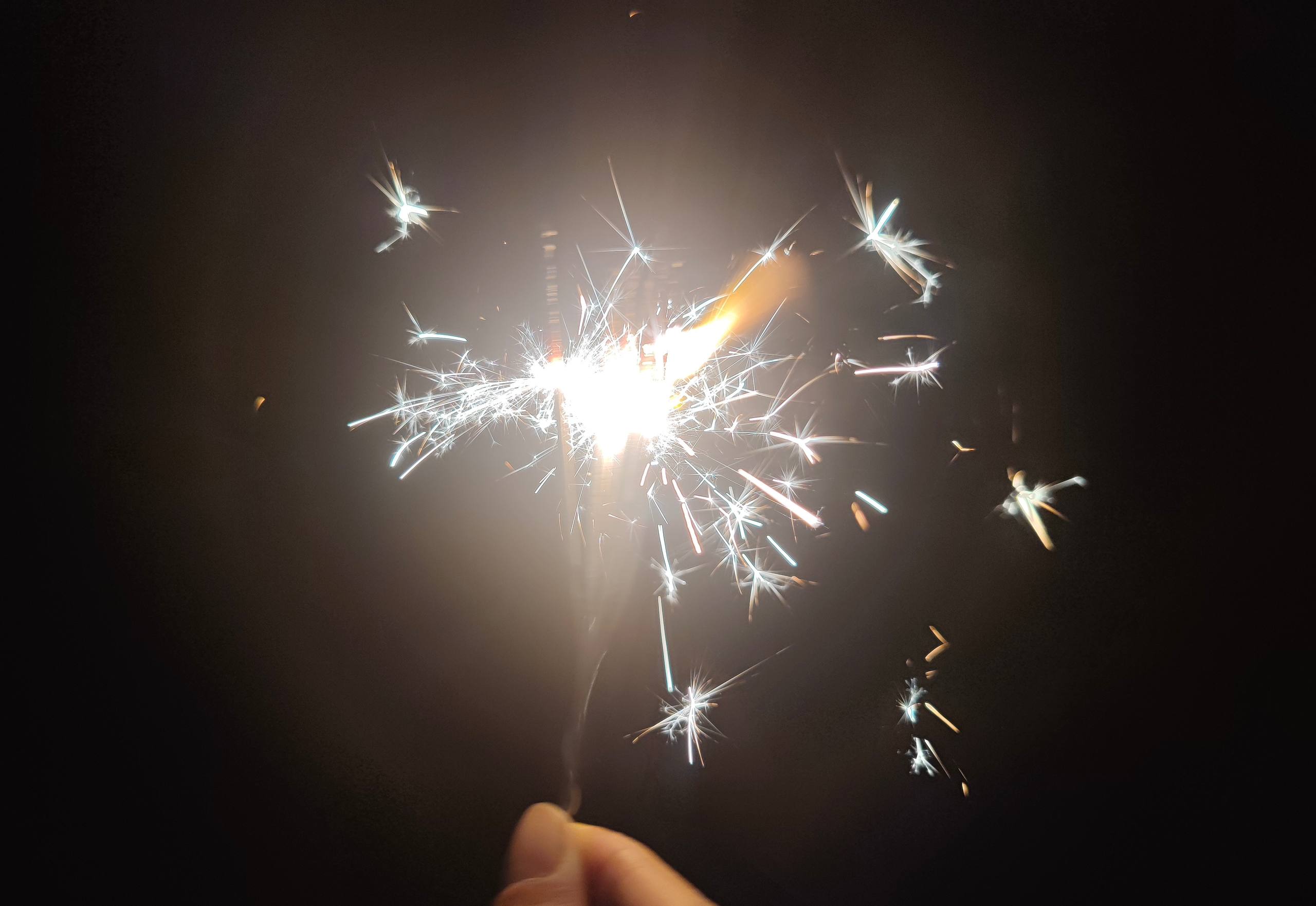 Hand holding a bright sparkler scattering sparks against a dark background.