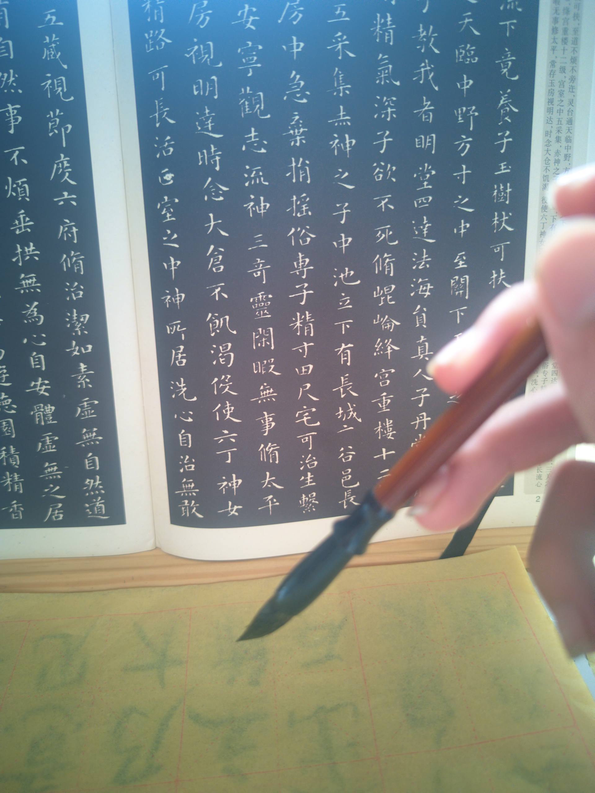Hand practicing Chinese calligraphy with a brush on gridded practice paper, using an open calligraphy copybook as a guide.