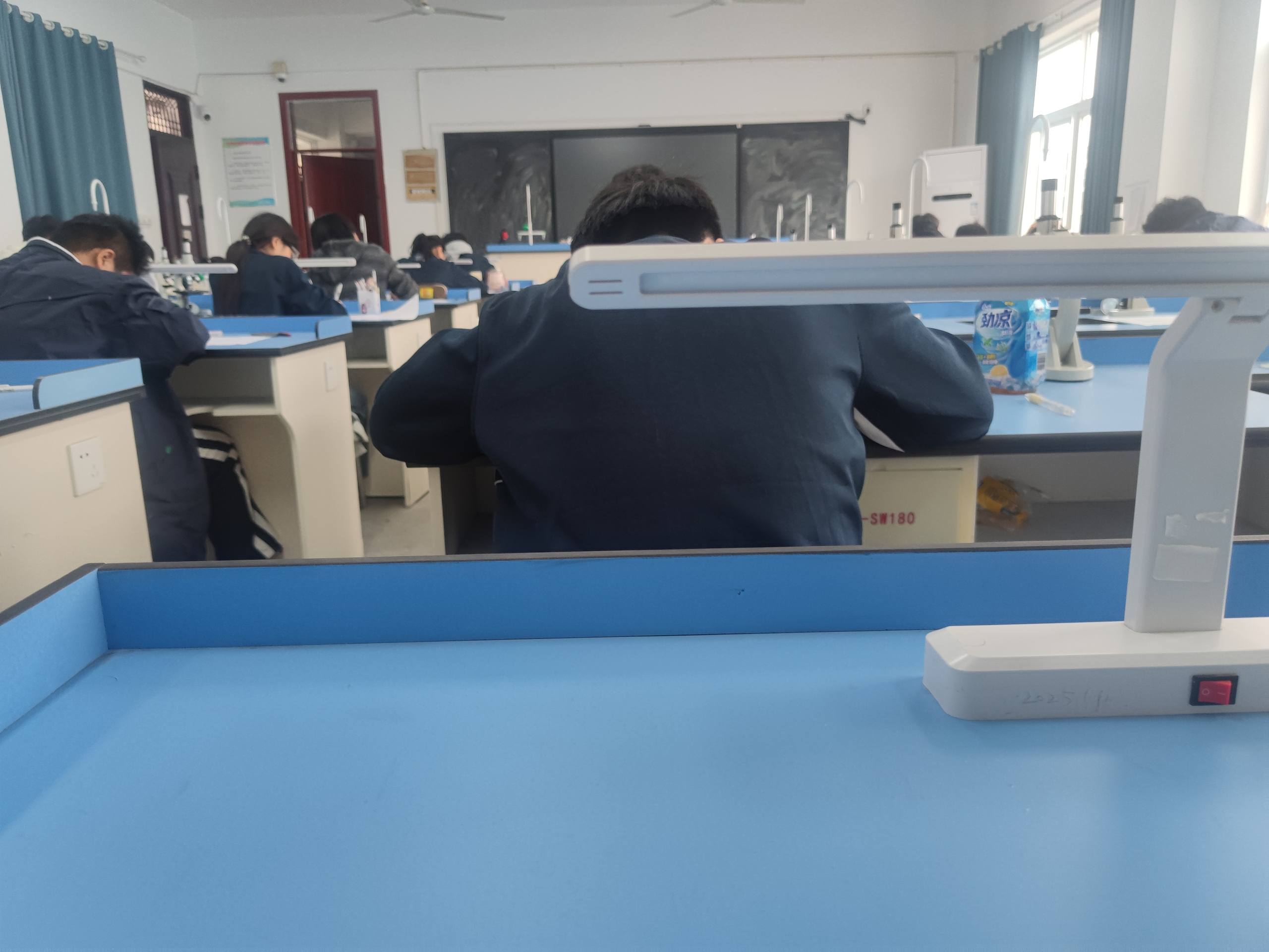 Students studying in a classroom or study room with blue desks, desk lamps, and a blackboard at the front.